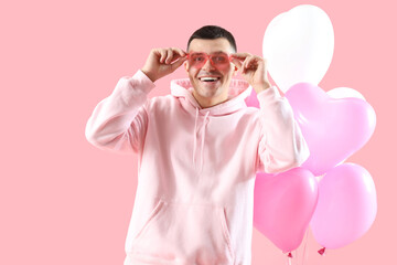 Wall Mural - Handsome man in heart-shaped sunglasses and balloons on pink background. Valentine's Day celebration