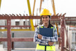 © Supachai - Asian woman engineer holding document smiling at construction site. Confident female Indian wearing protective helmet and vest working in factory making precast concrete wall for real estate housing.