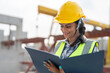 © Supachai - Asian woman engineer holding document smiling at construction site. Confident female Indian wearing protective helmet and vest working in factory making precast concrete wall for real estate housing.