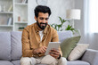 © Liubomir - Indian man smiles while browsing on a tablet, comfortably seated on a sofa in a well-lit modern apartment living room.