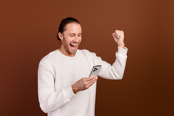 Wall Mural - Photo of excited lucky guy dressed white pullover rising fist winning game modern device isolated brown color background