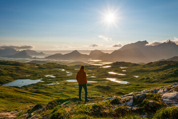 A man admiring the Lofoten mountains and nature from a high mountain top, from Justadtinden towards Leknes. Summer sun, green nature, hiking path.