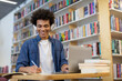 © Home-stock - Black student guy wearing headphones and taking notes in front of laptop in university library. Dedicated study session blending technology and traditional note-taking