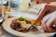 © pigwastudio - A person eating dinner with cutlery at a table covered with a tablecloth in a restaurant - close-up on hands and plate