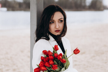 Wall Mural - Attractive brunette woman walking on the beach shore in moody cloudy windy weather with bouquet of red tulips flowers, dressed in white suit jacket. International Women`s Day 8th March concept
