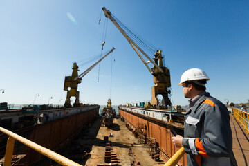 Professional overseeing extensive shipbuilding process, massive cranes erect vessel in dry dock against a clear sky. Maritime engineer in hardhat and reflective vest monitors construction.