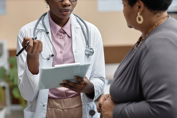 Cropped shot of Black woman general practitioner wearing lab coat with stethoscope questioning female patient while holding clipboard