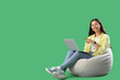 © Pixel-Shot - Young female programmer pointing at laptop while sitting on grey beanbag chair against green background