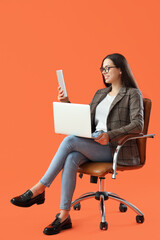 Pretty young female programmer with laptop and tablet computer sitting on armchair against orange background