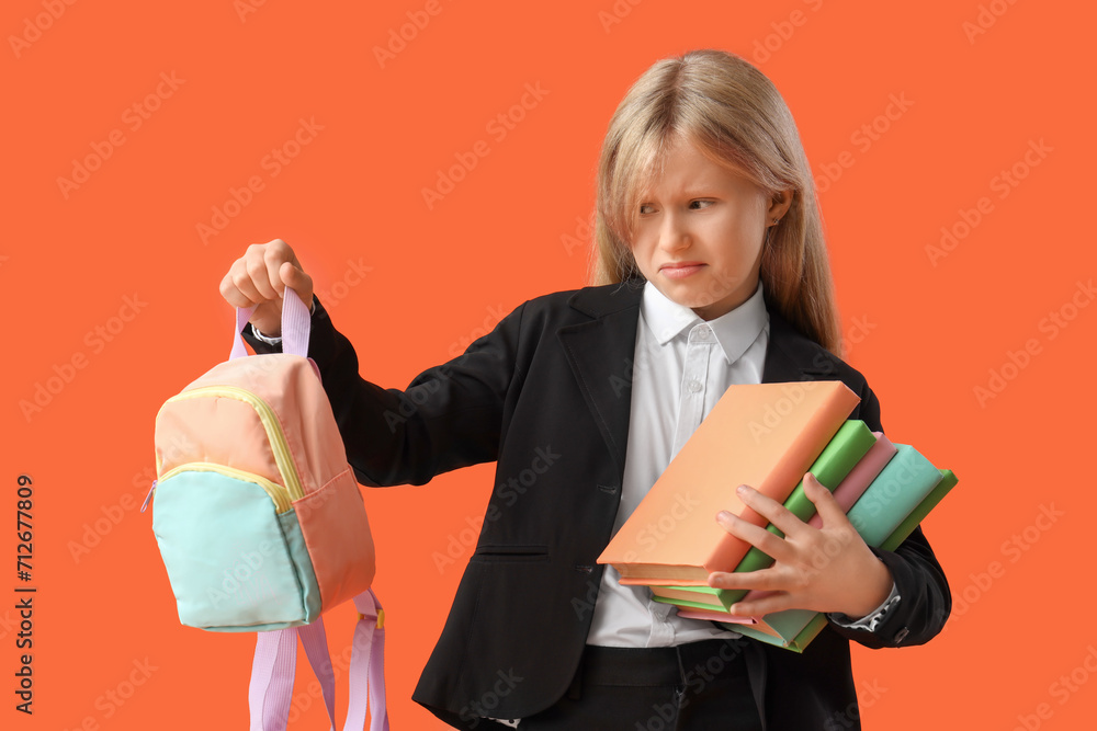 Upset little schoolgirl with small backpack and books on orange background