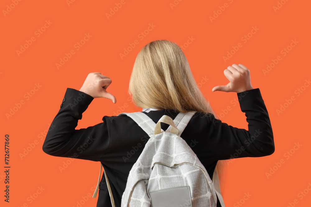 Little schoolgirl pointing at backpack on orange background, back view