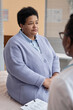 Vertical shot of unwell mature Black woman patient in light blue sweater sitting at examination table with unrecognizable health practitioner in hospital room