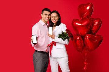 Wall Mural - Young couple with mobile phones, bouquet of roses and heart-shaped air balloons on red background. Online dating