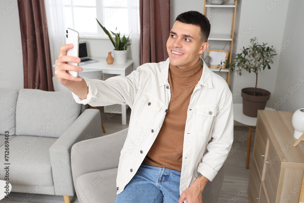 Young man with mobile phone taking selfie on armchair at home