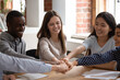 © fizkes - Smiling multiracial diverse millennial students sit at desk engaged in teambuilding activity at meeting in classroom, happy motivated multiethnic young people join fists hands show support and unity