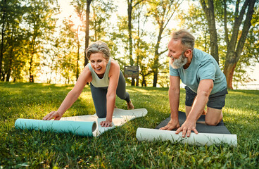 Wall Mural - Yoga practice on fresh air. Sporty aged man and woman in active attire chatting and smiling while putting two mats on grass during sunny morning weather. Concept of active and healthy retirement.