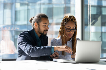 Internship. Confident black man mentor coach help teach explain work on project to biracial woman intern at workplace. Two young diverse mixed race colleagues share ideas interacting by laptop screen