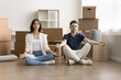 Calm young couple of new home owners meditating at paper moving boxes, sitting on floor, keeping zen hands and closed eyes, practicing stress relief after relocation activities