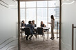 © fizkes - Candid shot through doorway of diverse business team and female leader discussing teamwork at large table. Group of young entrepreneurs negotiating, networking in modern office meeting room