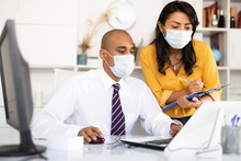 Portrait Of Two Office Employees In Medical Masks Concentrating On Work With Papers And Laptop. Necessary Precautions During Coronavirus Pandemic.