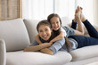 © fizkes - Little girl lying on moms back, smile look together at camera. Young woman enjoy carefree leisure lay on couch in cozy living room stare at cam feel happy. Custody, ties, motherhood, family relaxation