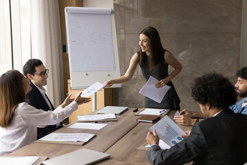 Wall Mural - Happy young project manager woman offering plan, management strategy to diverse team, giving paper marketing reports to colleagues, smiling. Business coach training employees for sales growth