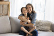 © fizkes - Loving mother sit on sofa cuddling tightly her little daughter, feeling pure love to child, spend priceless time together enjoy pastime at home. Cute girl sit on caring moms laps laugh look at camera