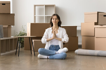 Peaceful young home owner woman meditating during stressful moving into new home, sitting on floor, keeping meditation pose, eyes closed, Namaste hand gestures with stacked boxes behind