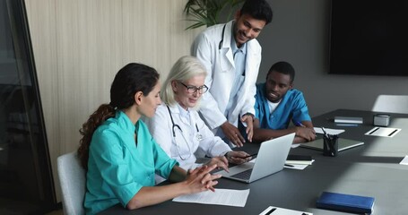 Wall Mural - Positive diverse team of younger doctors and elder boss, mentor meeting at laptop, talking, discussing professional medical application, online content, speaking together, pointing at screen