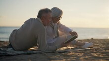 Senior Gray Haired Man Woman Reading Book Together Sitting Hugging On Sandy Sea Beach At Sunset. Happy Marriage, Old Family Spending Enjoy Time, Back View. Love Relationship, Resting Concept.