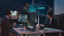 Medium Shot Of Black Male Cybersecurity Officer Pointing At Data Info On Display And Explaining Workflow Plan To Ethnically Diverse Colleagues While Co-working In Office Late At Night