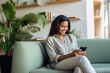 © AI_images - Happy young Latin woman sitting on sofa holding mobile phone using cellphone technology doing ecommerce shopping, buying online, texting messages.