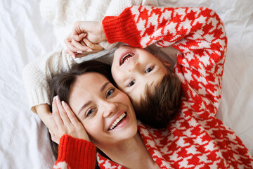 portrait of cheerful mother and toddler son in knitted sweaters looking at camera on bed at home, to