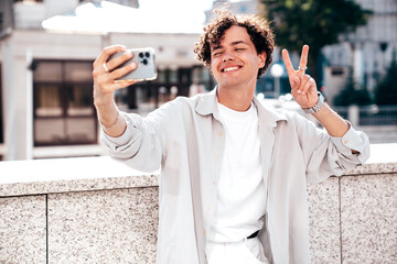 Wall Mural - Portrait of young attractive man with curly hair hairstyle. Smiling handsome male in casual stylish clothes posing in the street at sunny day. Cheerful and happy model outdoors. Take selfie photos