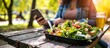 © Ilgun - Close up woman using meal tracker app on phone while eating salad at picnic table in the park on a break Healthy balanced diet lunch box Healthy diet plan for weight loss Selective focus
