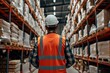 A warehouse worker, viewed from the back and clad in a white safety helmet and orange vest, standing in a big warehouse that full of shelves