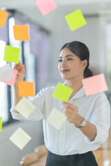 Wall Mural - Portrait of beautiful young woman standing in front of glass board and smiling on camera. Female office worker using laptop in company.