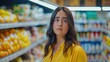 Young sad and uncertain latino woman retail buyer wearing bright yellow shirt with shelves with vegetables and other goods around her