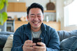 © Andrii  - Happy Relaxation: Asian Man Grinning with Phone on Sofa