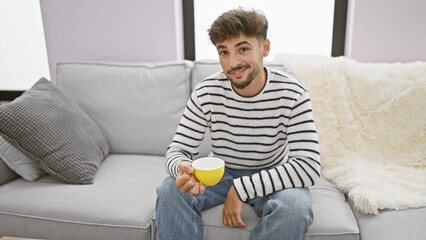 Canvas Print - Young, handsome arab man exuding confidence enjoying his morning cup of coffee, smiling joyfully while comfortably sitting on a sofa at home.