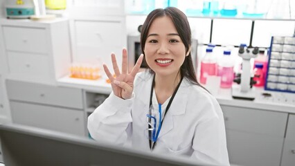 Canvas Print - Cheerful young chinese woman scientist in white lab coat showing number four with fingers, smiling and pointing up in the lab. a positive signal in the world of pharmacy!