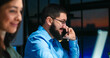 © ihorvsn - Caucasian handsome man in glasses and beard talking on mobile phone and smiling late in evening. Blurred female colleague face. Male office worker speaking on cellphone. Night shift job.