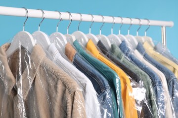 Poster - Dry-cleaning service. Many different clothes in plastic bags hanging on rack against light blue background, closeup