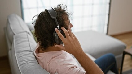 Sticker - A relaxed man listens to music on headphones while lounging on a sofa in a bright, modern living room.