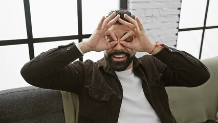 Poster - Hilarious young man at home making crazy binoculars look with 'ok' finger gesture, cheerfully sticking his tongue out, eyes gleaming through fingers.