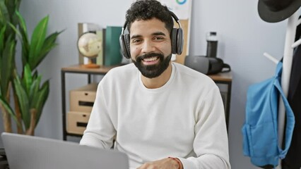 Sticker - Handsome hispanic man with beard wearing headphones smiles at camera in modern office with plant and globe.