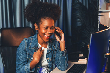 Young African creator calling with smartphone and looking on screen monitor laptop, wearing jeans shirt. Getting project or study new technology at workplace. Theme of teenager winning. Tastemaker.