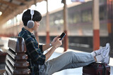 Young man in headphones using mobile phone while sitting on bench at railway station