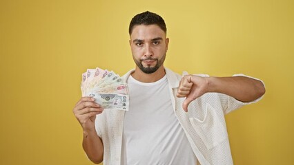 Poster - Unhappy young arab man showing a thumbs down sign with united arab emirates banknotes in hand on a yellow background, symbol of rejection and dislike