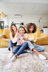 Wall Mural - Vertical photo three young multi-ethnic women playing video games together at home. Group girls enjoying free time with popcorn in community in living room. Generation z and positive friendships.